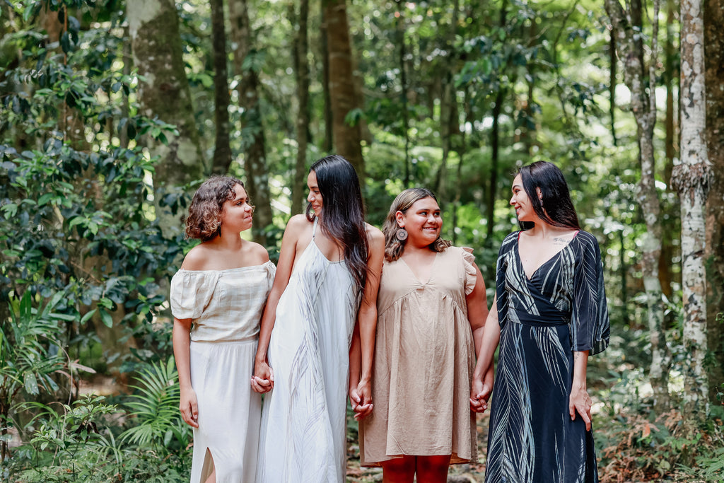 Four young Indigenous women holding hands in nature, wearing earthy coloured dresses 